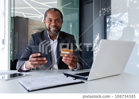 Senior African American man in suit sitting at desk in office. He is holding a credit card and a phone. Successfully paid online purchases, orders, bills. Happy looking at camera. Senior African American man in suit sitting at desk in office. He is holding a credit card and a phone. Successfully paid online purchases, orders, bills. Happy looking at camera. 108734385