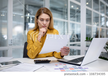 Upset blonde businesswoman working in a modern office, sitting at a desk in front of a notebook, thoughtfully working with business papers, carefully reviewing important information. 108734395