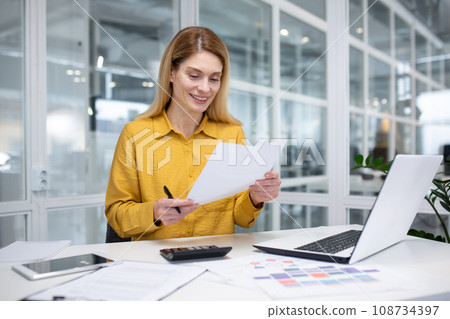 An attractive blonde woman works in a modern office, sits at a desk in front of a notebook, does paperwork, looks intently at the computer screen, jots down information. An attractive blonde woman works in a modern office, sits at a desk in front of a notebook, does paperwork, looks intently at the computer screen, jots down information. 108734397