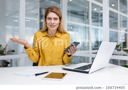 Dissatisfied blonde woman businesswoman sitting at desk in bright office, holding smartphone in hand, losing the lottery, reading bad news, angry because of non-working phone, looking at the camera. 108734405