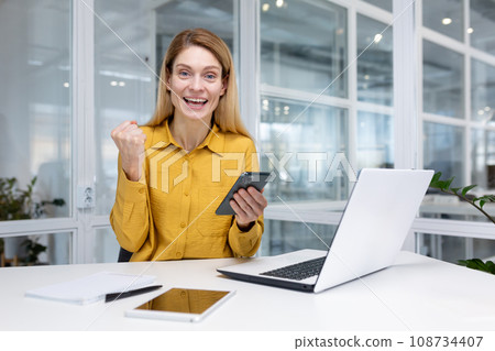 Happy blonde business woman sitting at a desk in a bright office, holding a smartphone in her hand, enjoying winning the lottery, reading good news, getting a promotion, looking at camera. Happy blonde business woman sitting at a desk in a bright office, holding a smartphone in her hand, enjoying winning the lottery, reading good news, getting a promotion, looking at camera. 108734407