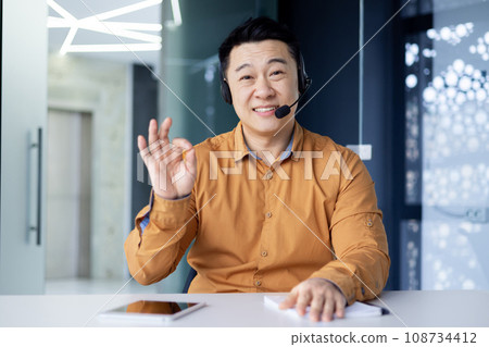 Happy asian office worker sitting at desk in bright office wearing headset and talking on video call, chinese businessman smiling and showing ok sign looking at camera, camera view. 108734412