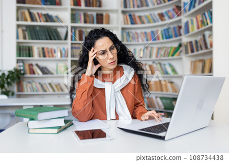 Tired young hispanic female teacher sitting at desk in front of laptop in school room holding hand on head, overwork in office headache, online learning concept. 108734438