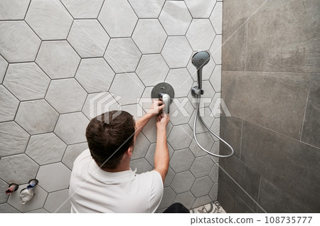 Back view of male plumber installing wall mounted shower system in apartment under renovation. Man repairing metal shower faucet handle while working on bathroom renovation. Plumbing works concept. Back view of male plumber installing wall mounted shower system in apartment under renovation. Man repairing metal shower faucet handle while working on bathroom renovation. Plumbing works concept. 108735777