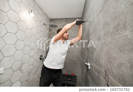Side view of male worker using electric drilling machine while working on bathroom renovation. Man drilling wall with cordless power drill while installing shower system at home. Side view of male worker using electric drilling machine while working on bathroom renovation. Man drilling wall with cordless power drill while installing shower system at home. 108735782