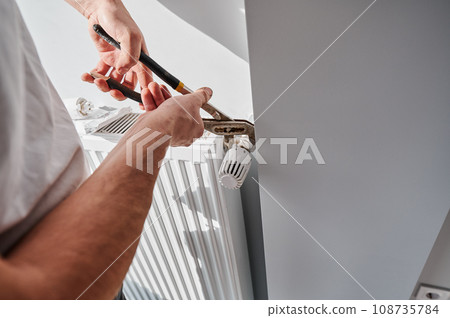 Close up of man using wrench tool while installing heating radiator in apartment under renovation. Male plumber working on heating system installation. Home renovation and plumbing works concept. Close up of man using wrench tool while installing heating radiator in apartment under renovation. Male plumber working on heating system installation. Home renovation and plumbing works concept. 108735784