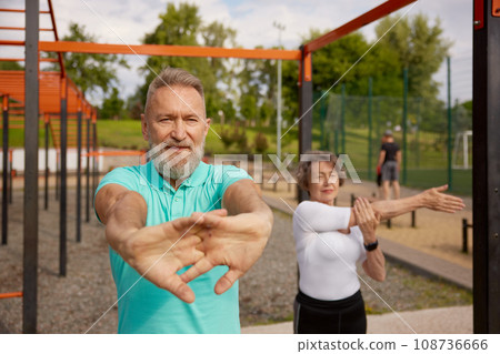 Seniors couple stretching arms before exercise at training field outdoors Seniors couple stretching arms before exercise at training field outdoors 108736666