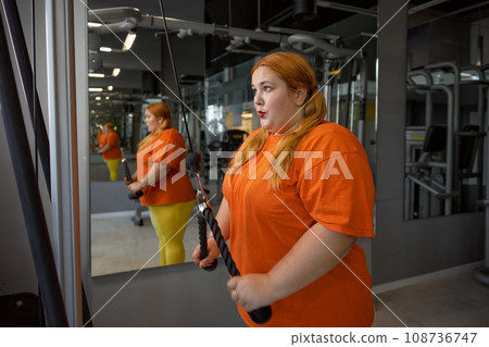 Exhausted excess overweight woman training with equipment at gym 108736747