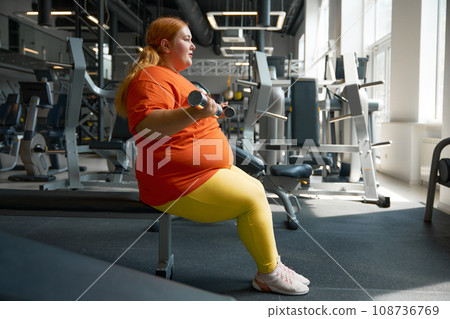 Plus size woman doing exercise on biceps training with dumbbells at gym Plus size woman doing exercise on biceps training with dumbbells at gym 108736769