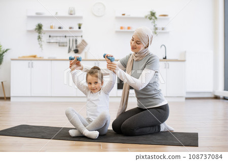 Woman in hijab helping sweet daughter with lifting dumbbells on black mat in kitchen. Woman in hijab helping sweet daughter with lifting dumbbells on black mat in kitchen. 108737084