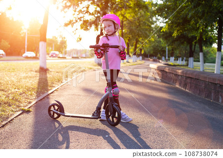 a beautiful girl in a pink helmet and protective equipment stands with a scooter on an alley in the city against the backdrop of a sunny sunset in summer. 108738074