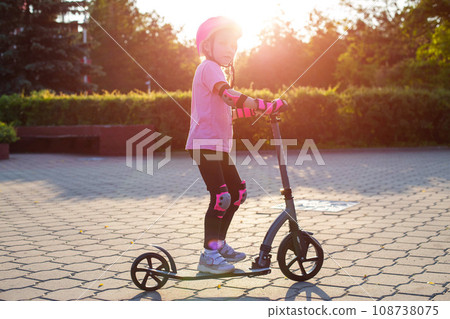 A beautiful caucasian girl of seven years old rides a scooter in a pink outfit, a helmet and knee pads on a sunny summer day. Safety in active sports. A beautiful caucasian girl of seven years old rides a scooter in a pink outfit, a helmet and knee pads on a sunny summer day. Safety in active sports. 108738075