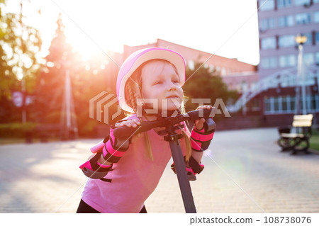 A beautiful caucasian girl of seven years old in a pink protective helmet and outfit is fooling around on a scooter in the summer. Smiling girl laughs next to the scooter. Sunset A beautiful caucasian girl of seven years old in a pink protective helmet and outfit is fooling around on a scooter in the summer. Smiling girl laughs next to the scooter. Sunset 108738076