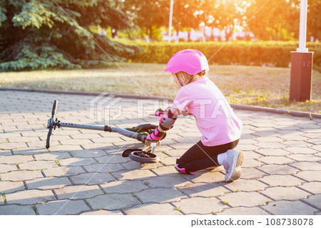 A 7-year-old girl in a pink helmet and protective gear fell off a scooter. The concept of safe riding a scooter and a bicycle without injury. Copy space for text A 7-year-old girl in a pink helmet and protective gear fell off a scooter. The concept of safe riding a scooter and a bicycle without injury. Copy space for text 108738078