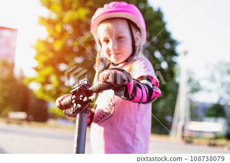 A beautiful caucasian girl of 7 years old in a pink helmet and armlets stands on a scooter. Summer sunny day. A beautiful caucasian girl of 7 years old in a pink helmet and armlets stands on a scooter. Summer sunny day. 108738079