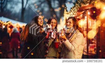 Happy young women attending winter fair in evening, drinking hot drinks, eating hot dogs. Merry season and cheerful time 108738181