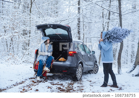 In a winter forest, a woman sits in the trunk of a car, and a man carries a Christmas tree In a winter forest, a woman sits in the trunk of a car, and a man carries a Christmas tree 108738755