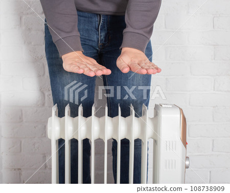 A man warms his hands on an electric oil heater at home. The concept of cold in apartments in autumn and winter. Turn on the heating. 108738909