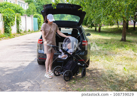A beautiful girl puts a baby stroller into the open trunk of a car. Family car concept. Sunny weather in summer. A beautiful girl puts a baby stroller into the open trunk of a car. Family car concept. Sunny weather in summer. 108739029