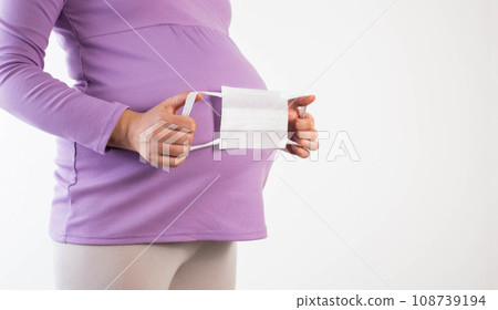 A pregnant girl in a lilac blouse holds a protective medical mask against the background of her belly. The concept of protection against colds infectious diseases coronavirus and swine flu. Copy space A pregnant girl in a lilac blouse holds a protective medical mask against the background of her belly. The concept of protection against colds infectious diseases coronavirus and swine flu. Copy space 108739194