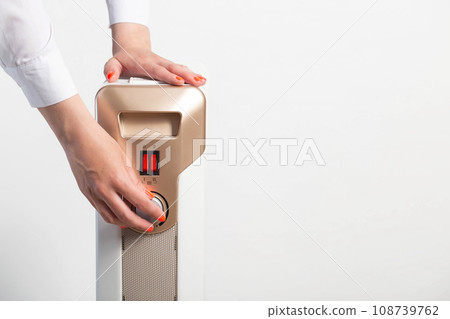 A woman s hand with a beautiful manicure regulates the room heating temperature on an electric heater. The concept of turning on the heating in the autumn-winter period. Cold in apartments, close-up. 108739762