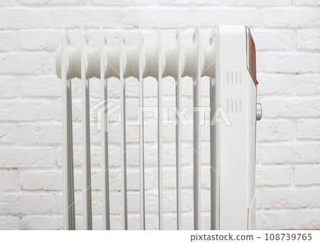 Modern electric oil heating radiator against a white brick wall. The concept of turning on heating in apartments. The cold season is autumn and winter. Modern electric oil heating radiator against a white brick wall. The concept of turning on heating in apartments. The cold season is autumn and winter. 108739765