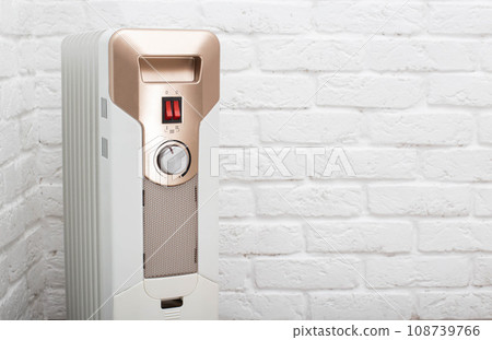 Electric heating radiator with temperature control on the background of a white brick wall. The concept of heating a room without turning on the heating in the apartment. The cold season is autumn and Electric heating radiator with temperature control on the background of a white brick wall. The concept of heating a room without turning on the heating in the apartment. The cold season is autumn and 108739766