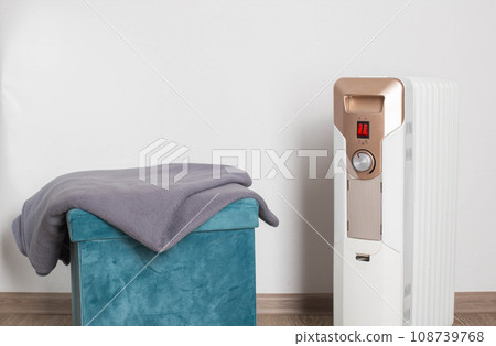A gray blanket lies on an ottoman next to an electric heater. The concept of heating a room without heating in an apartment. Copy space for text A gray blanket lies on an ottoman next to an electric heater. The concept of heating a room without heating in an apartment. Copy space for text 108739768