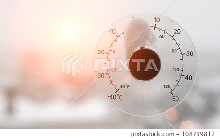 The rays of the setting sun in the sky against the background of a street thermometer with a snow cap on the window. Snowy weather and warming concept. Weather forecast Copy space for text The rays of the setting sun in the sky against the background of a street thermometer with a snow cap on the window. Snowy weather and warming concept. Weather forecast Copy space for text 108739812