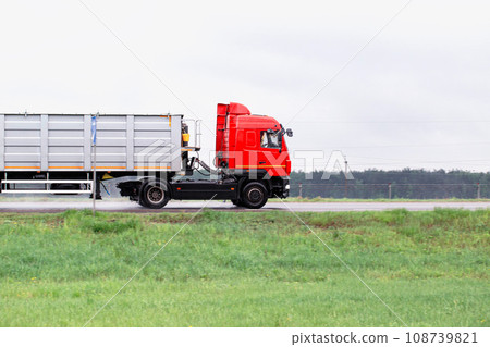 A red truck with a semi-trailer transports bulk cargo along the road in cloudy weather. Side view. Copy space for text, industry 108739821