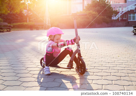 A beautiful caucasian girl of seven years old in a pink protective helmet and outfit is fooling around on a scooter in the summer. Smiling girl laughs next to the scooter. Sunset A beautiful caucasian girl of seven years old in a pink protective helmet and outfit is fooling around on a scooter in the summer. Smiling girl laughs next to the scooter. Sunset 108740134