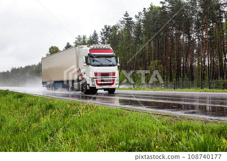 A beautiful truck with a semi-trailer transports cargo in rainy weather on a slippery road against the background of a forest. Freight transportation market, import substitution, business, trucking A beautiful truck with a semi-trailer transports cargo in rainy weather on a slippery road against the background of a forest. Freight transportation market, import substitution, business, trucking 108740177