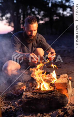 A young man with a beard prepares a dish of bacon with scrambled eggs on a fire. Cooking and outdoor recreation in summer. A young man with a beard prepares a dish of bacon with scrambled eggs on a fire. Cooking and outdoor recreation in summer. 108740935