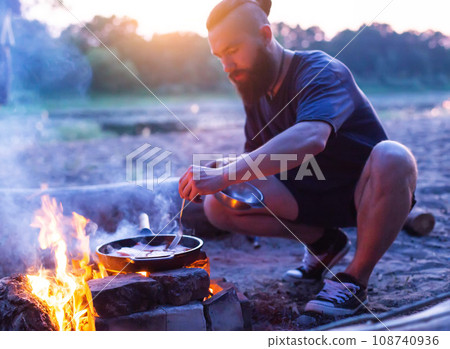 A brutal man with a beard cooks food in a frying pan on a fire in nature. Cooking bacon on the river bank. Camping. A brutal man with a beard cooks food in a frying pan on a fire in nature. Cooking bacon on the river bank. Camping. 108740936