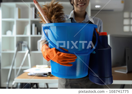 Young woman holding bucket with cleaning tools and equipment while standing in office 108741775