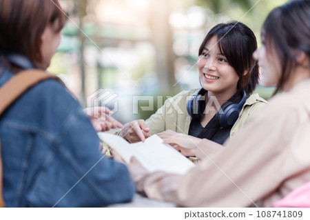 Group of young Asian college students sitting on a bench in a campus relaxation area, talking, sharing ideas, doing homework or tutoring for the exam together Group of young Asian college students sitting on a bench in a campus relaxation area, talking, sharing ideas, doing homework or tutoring for the exam together 108741809