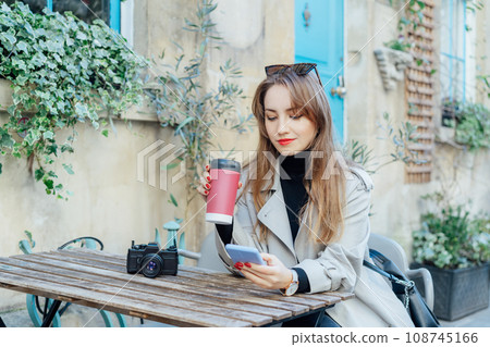 Female traveler chatting, sharing files, resting in cafe terrace table with vintage camera and coffee in reusable cup after city walk. Girl using internet in wifi zone, enjoying coffee and leisure Female traveler chatting, sharing files, resting in cafe terrace table with vintage camera and coffee in reusable cup after city walk. Girl using internet in wifi zone, enjoying coffee and leisure 108745166