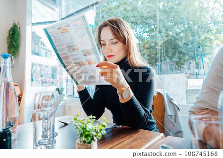Young woman reading menu sitting with friend at casual cafe. Smiling woman checking menu, choosing meal or drink, making order. Try new foods while traveling. real people. copy space. Selective focus. Young woman reading menu sitting with friend at casual cafe. Smiling woman checking menu, choosing meal or drink, making order. Try new foods while traveling. real people. copy space. Selective focus. 108745168