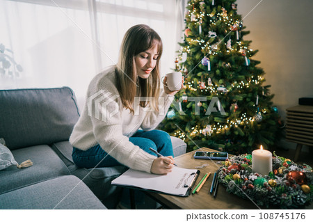 Young Woman with cup is hand writing New year's goals on note paper in new year day. New year resolutions list. Goals, resolutions, plans, action, checklist concept. New Year template, copy space Young Woman with cup is hand writing New year's goals on note paper in new year day. New year resolutions list. Goals, resolutions, plans, action, checklist concept. New Year template, copy space 108745176