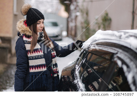 A woman is cleaning snowy window on a car with snow scraper. Pretty woman warmy dressed clean her car outdoors. Cold snowy and frosty morning. Black car A woman is cleaning snowy window on a car with snow scraper. Pretty woman warmy dressed clean her car outdoors. Cold snowy and frosty morning. Black car 108745897