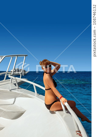 A beautiful blonde woman in a black bikini and white shirt is enjoying her summer vacation sunbathing on a luxury yacht A beautiful blonde woman in a black bikini and white shirt is enjoying her summer vacation sunbathing on a luxury yacht 108746132
