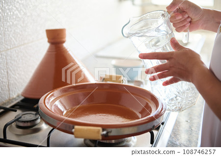 Closeup housewife beginning cooking meal in tajine clay pot, pouring some water inside a clay dish standing on a stove Closeup housewife beginning cooking meal in tajine clay pot, pouring some water inside a clay dish standing on a stove 108746257