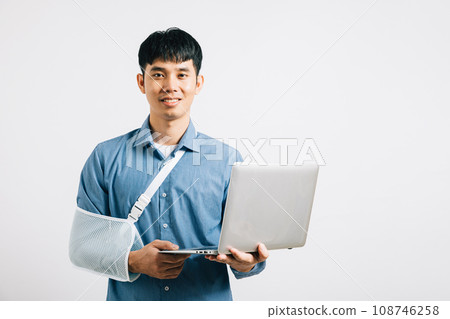 Injured Asian businessman, with a bandaged broken arm, remains resilient, working on a laptop. Studio shot isolated on white, symbolizing commitment to work and recovery. Injured Asian businessman, with a bandaged broken arm, remains resilient, working on a laptop. Studio shot isolated on white, symbolizing commitment to work and recovery. 108746258