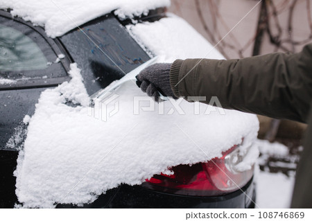 First person view of man cleaning snowy window on a car with snow scraper. Focus on the scraper. Cold snowy and frosty morning. Black car 108746869