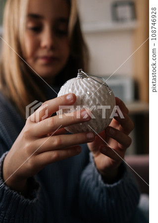 Close-up vertical portrait of adorable little girl holding and looking festive Christmas ball with happy expression. Smiling kid decorates xmas tree for New Year at home in dark living room alone. 108746928