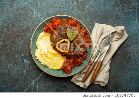 Traditional Italian dish Ossobuco all Milanese made with cut veal shank meat with vegetable tomato sauce served with corn polenta on ceramic plate top view on rustic stone background Traditional Italian dish Ossobuco all Milanese made with cut veal shank meat with vegetable tomato sauce served with corn polenta on ceramic plate top view on rustic stone background 108747705