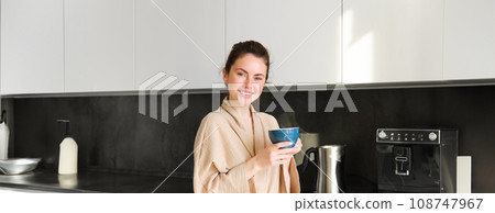 Daily routine and lifestyle. Young beautiful woman in bathrobe, standing in kitchen with cup of coffee, drinking tea, smiling and looking happy 108747967