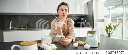 Attractive young cheerful girl baking at the kitchen, making dough, holding recipe book, having ideas 108747971