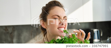 Funny and cute brunette woman bites delicious, fresh green parsley, eating herbs, cooking salad in kitchen Funny and cute brunette woman bites delicious, fresh green parsley, eating herbs, cooking salad in kitchen 108747991