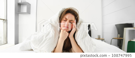 Portrait of woman rolled in blankets in bed, frowning and feeling unwell, has hangover, headache in morning, resting at home Portrait of woman rolled in blankets in bed, frowning and feeling unwell, has hangover, headache in morning, resting at home 108747995